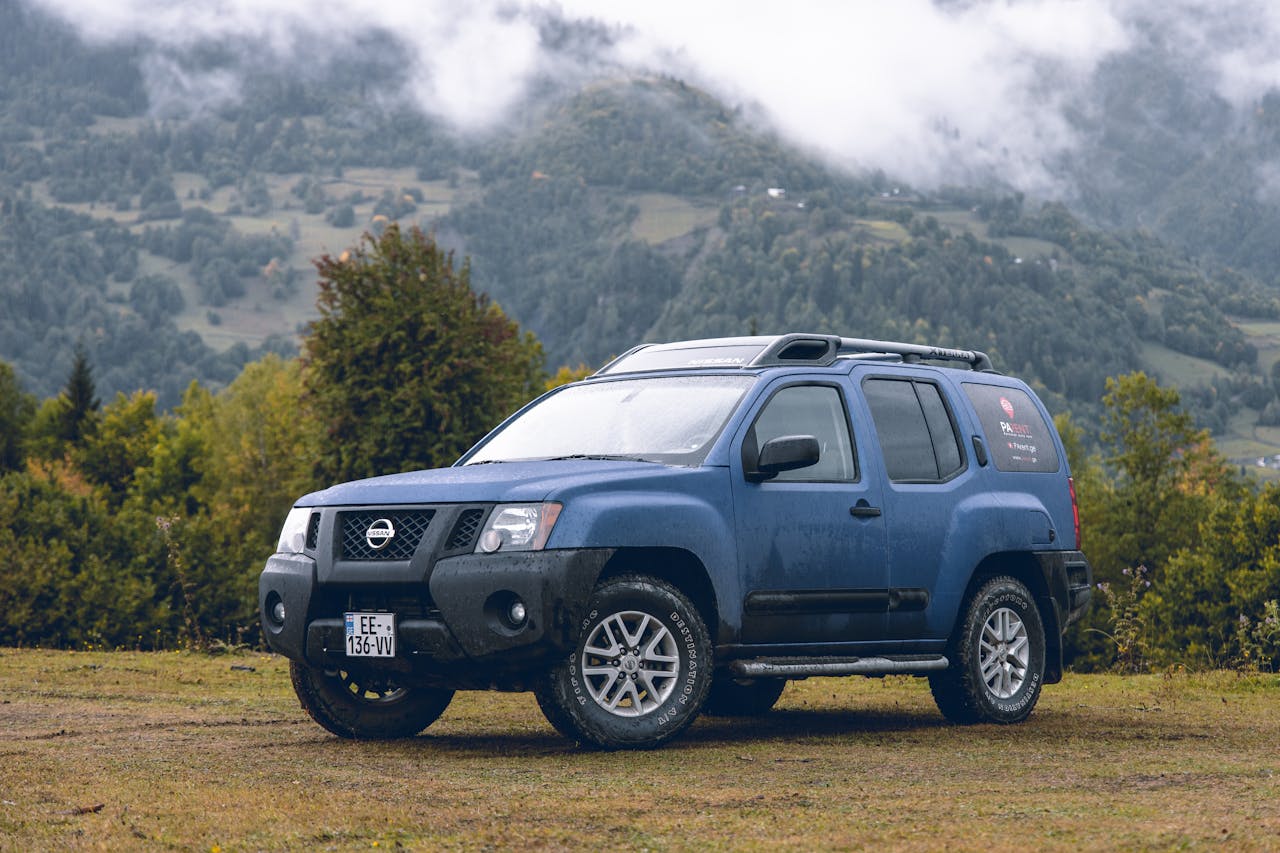 A rugged blue Nissan SUV parked against a picturesque mountain backdrop with lush greenery and misty clouds.
